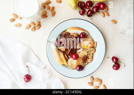 Trending Food Cerealien Pfannkuchen. Kleine Pfannkuchen mit frischen Birnen, Kirschen, Mandeln in einem Metall blauen Teller auf dem Hintergrund der Küchentisch mit Fr. Stockfoto