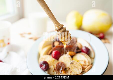 Trending Food Cerealien Pfannkuchen. Honig tropft aus einem Holzstab auf kleine Pfannkuchen mit Birnen, Kirschen, Nüsse in Metall blauen Teller vor dem Hintergrund Stockfoto