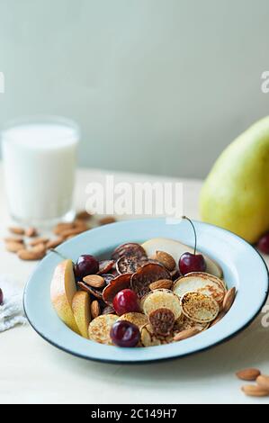 Kleine Pfannkuchen mit Birnen, Kirschen, Nüsse in Metallplatte vor dem Hintergrund der Küchentisch mit Obst und Milch. Trending Lebensmittel Müsli Pfannkuchen. br Stockfoto