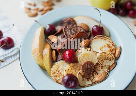 Winzige Pfannkuchen mit Birnen, Kirschen, Nüsse in Metallplatte vor dem Hintergrund der Küchentisch mit Obst. Trending Lebensmittel Müsli Pfannkuchen. Hell und Stockfoto