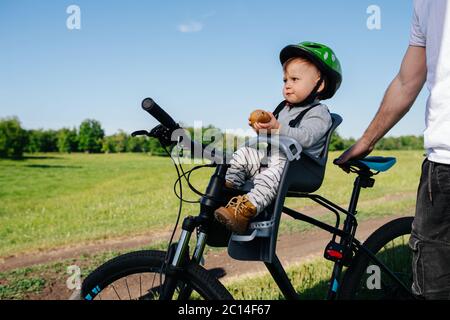 Mürriges Baby sitzt auf einem Sicherheitsstuhl auf einem Fahrrad, während sein Vater ihn hält Stockfoto