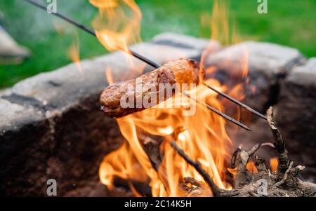 Wurst stach auf eine Eiserne Gabel und toastete über auf Lagerfeuer in der Natur. Würstchen backen. Nahaufnahme auf Feuer und auf gebratene Wurst. Stockfoto