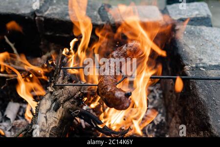 Wurst stach auf eine Eiserne Gabel und toastete über auf Lagerfeuer in der Natur. Würstchen backen. Nahaufnahme auf Feuer und auf gebratene Wurst. Stockfoto