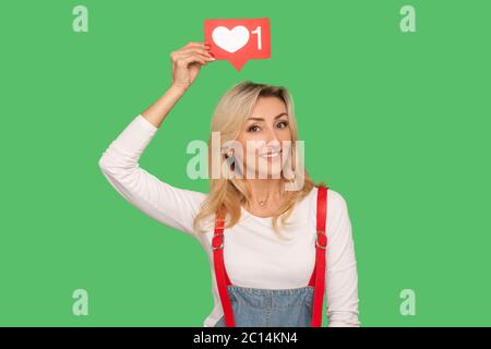 Folgen und mögen Social Media Blog. Portrait von schönen lächelnden erwachsenen Frau in stilvollen Denim Overalls halten Netzwerk Herz Symbol über Kopf, emoji novi Stockfoto