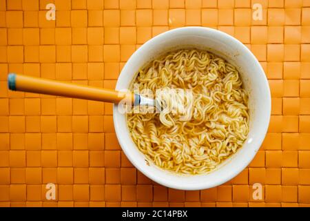 Hand eines Mannes, der eine Gabel mit gekochten Instant-Nudeln aus einer Keramikschale auf orangefarbenem Hintergrund hält. Draufsicht. Billiges Essen Stockfoto