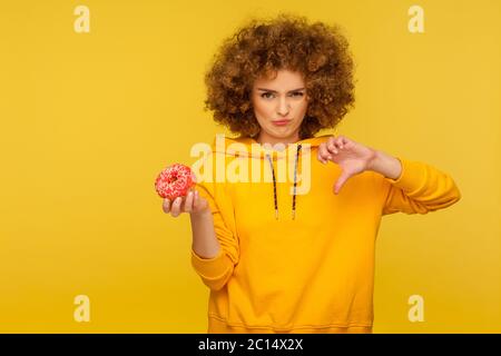 Abneigung gegen Junk Food, zuckerhaltige Gebäck. Portrait der unzufriedenen unglücklich lockig-haarige Frau im urbanen Stil Hoodie zeigt Daumen nach unten und hält süß d Stockfoto