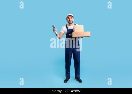 Volle Länge überrascht Lieferung Mann in Uniform und Handschuhe halten Handy, Kartons und Blick auf die Kamera mit staunenden Ausdruck. Kurier e Stockfoto