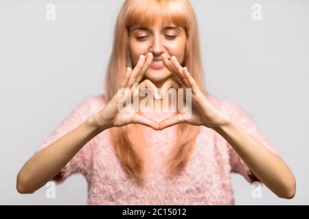 Studio Portrait von freundlich lächelnden Teenager Frau, die eine Herzbewegung mit ihren Fingern macht, zeigt ihre Liebe, konzentriert sich auf die Hände, isoliert auf grauem Hintergrund. Stockfoto