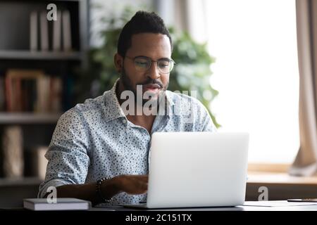 Afrikanischer Mann, der am Schreibtisch sitzt und einen Laptop benutzt, um fernzuarbeiten Stockfoto