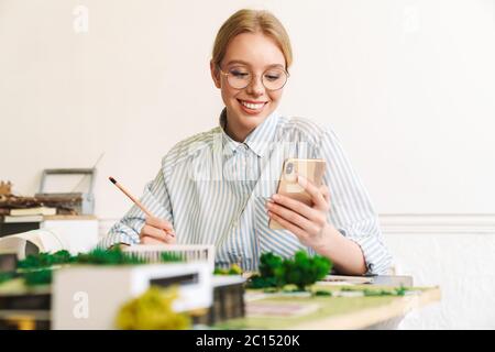 Foto von fröhlichen jungen Frau Architekt mit Handy beim Entwurf mit Haus Modell am Arbeitsplatz Stockfoto