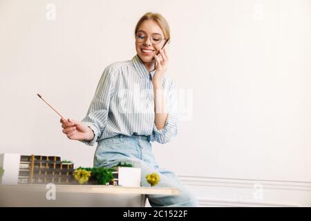 Foto von fröhlichen jungen Frau Architekt im Gespräch auf Handy während der Entwurf mit Haus Modell am Arbeitsplatz zu entwerfen Stockfoto