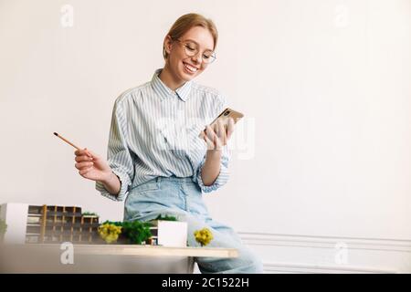 Foto von fröhlichen jungen Frau Architekt mit Handy beim Entwurf mit Haus Modell am Arbeitsplatz Stockfoto