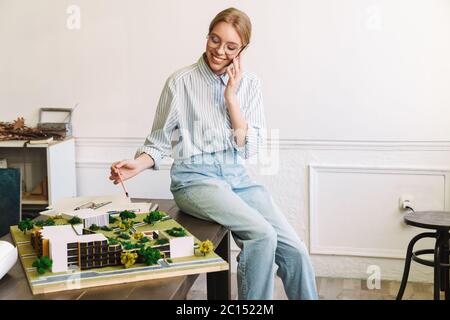 Foto von fröhlichen jungen Frau Architekt im Gespräch auf Handy während der Entwurf mit Haus Modell am Arbeitsplatz zu entwerfen Stockfoto