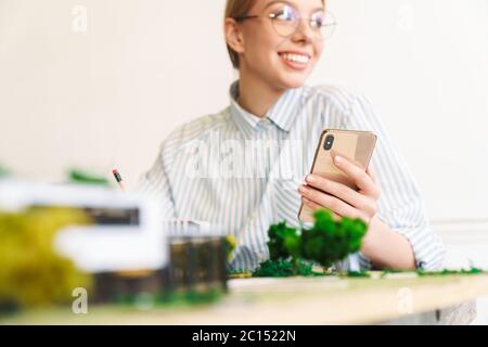 Foto von fröhlichen jungen Frau Architekt mit Handy beim Entwurf mit Haus Modell am Arbeitsplatz Stockfoto