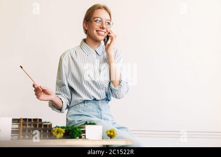 Foto von fröhlichen jungen Frau Architekt im Gespräch auf Handy während der Entwurf mit Haus Modell am Arbeitsplatz zu entwerfen Stockfoto