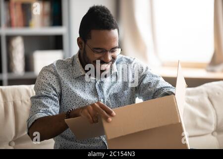 Afrikanischer Mann, der das Paket öffnet, sitzt auf der Couch zu Hause Stockfoto