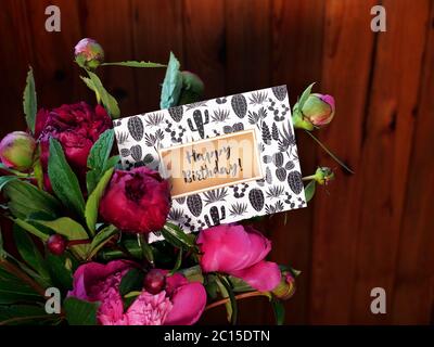 Nahaufnahme von rosa Pfingstrosen Blumen und Vase auf Holzhintergrund mit Happy Birthday Karte. Das Foto wurde mit natürlichem Tageslicht aufgenommen. Stockfoto