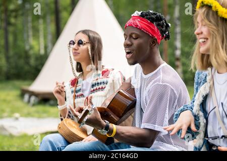 Talentierter junger schwarzer Kerl, der Gitarre spielt und singt, während er mit engen Freunden eine Campingparty genießt Stockfoto