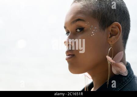 Nahaufnahme einer jungen Afro-Amerikanerin mit Stern-Glitzer um das Auge, die lange Ohrringe mit rosa Leder im Freien trägt Stockfoto