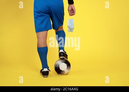 Fußballspieler mit Gesichtsmaske in der Hand wegen covid19 Coronavirus Pandemie und Ball im Fuß auf gelbem Hintergrund Stockfoto