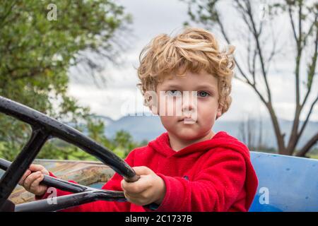 Portrait eines jungen, jungen, blonden Jungen, der auf einem Traktor sitzt und das Lenkrad hält Stockfoto