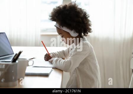 Afrikanisches Mädchen trägt Kopfhörer und macht Hausaufgaben am Schreibtisch Stockfoto