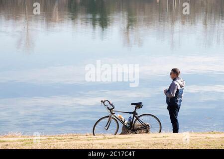 Belgrad, Serbien - 17. Februar 2020: Ein junger Mann steht allein neben seinem Fahrrad auf einem Seeufer, mit Reflexionen auf dem Wasser Stockfoto
