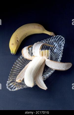 Banane in Glas geschält Banane Split antike Schüssel mit selektivem Fokus und eine ungeschält auf schwarzem Rücken Stockfoto