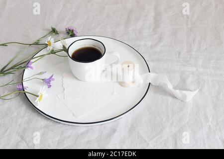 Frühling, Sommer Stillleben Szene. Unbeschriftete Grußkarten-Mockups aus Baumwollpapier auf Keramikplatte. Tasse Kaffee, Seidenband, Gänseblümchen und Bluebells Blumen Stockfoto