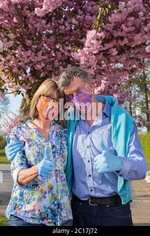 Seniorenpaar mit einer positiven Einstellung während der Pandemie Covid-19 Arm in Arm vor rosa Frühlingsblüten stehend, mit bunten Masken und Glo Stockfoto