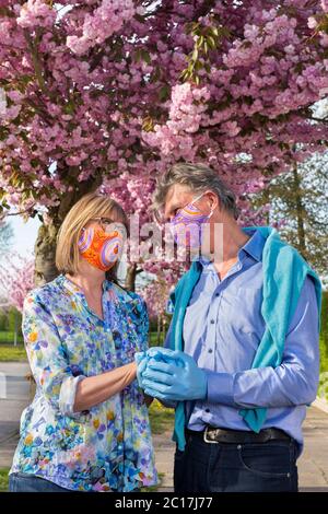 Glückliches Seniorenpaar tief verliebt vor rosa Frühlingsblüte stehend mit Händen in Gesichtsmasken und Handschuhen während der Covid-19 coronaviru Stockfoto