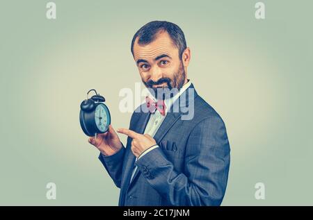 Ein reifer Mann im Anzug zeigt einen Wecker. Geschäftsmann mit Wecker in der Hand und Blick auf die Kamera. Mischrasse bärtige Modell isoliert auf grünen yel Stockfoto