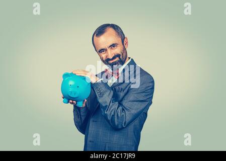 Sly junger Mann hält Sparschwein Verschwörung. Bärtiger Hipster Geschäftsmann isoliert auf grün gelb Studio Wand Hintergrund. Negativer Gesichtsausdruck, huma Stockfoto