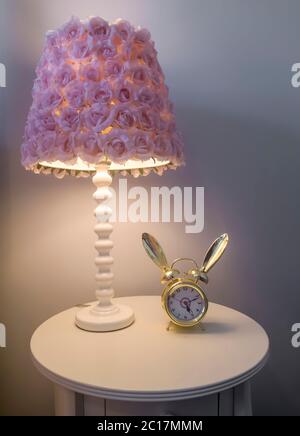 Lampenschirm mit violetten Blumen neben einem Wecker auf dem runden Beistelltisch Stockfoto