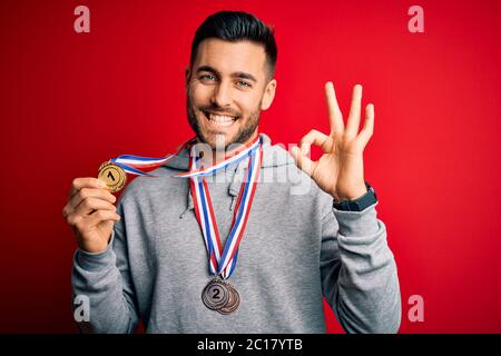 Junge gut aussehende erfolgreiche Mann Medaillen über isolierten roten Hintergrund zu tun ok Zeichen mit Fingern, ausgezeichnetes Symbol Stockfoto