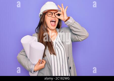 Junge hispanische Architektin Frau trägt Baumeister Hut und hält Blaupausen über gelben Hintergrund mit glücklichen Gesicht lächelnd tun ok Zeichen mit Hand auf Ey Stockfoto