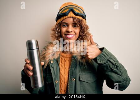 Junge afroamerikanische Skifahrerin Frau trägt Skibrille trinken Thermo mit Kaffee glücklich mit großen Lächeln tun ok Zeichen, Daumen hoch mit den Fingern, excellen Stockfoto