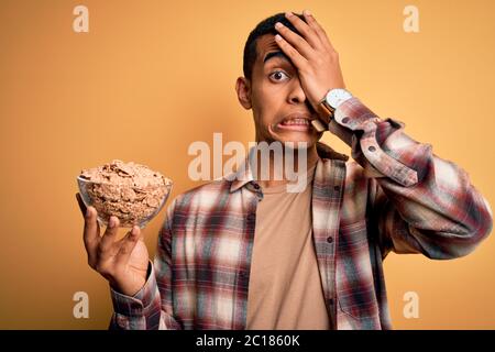 Junge gutaussehende afroamerikanische Mann hält Schüssel mit gesunden Cornflakes Getreide mit Hand auf Kopf gestresst, schockiert mit Scham und Überraschung Gesicht, ang Stockfoto