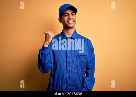 Junge afroamerikanische Mechaniker Mann trägt blaue Uniform und Kappe über gelbem Hintergrund lächelnd mit glücklichem Gesicht suchen und zeigen auf die Seite mit Stockfoto