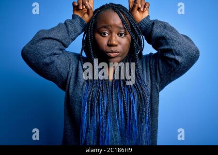 afroamerikanische plus size Frau mit Zöpfen tragen lässigen Pullover über blauem Hintergrund tun lustige Geste mit Finger über Kopf als Stierhörner Stockfoto