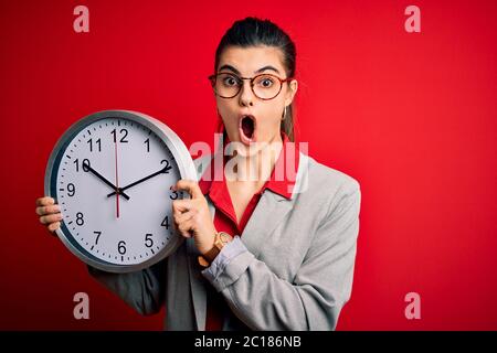 Junge schöne Brünette Geschäftsfrau tun Countdown hält große Uhr erschrocken im Schock mit einem Überraschungsgesicht, Angst und aufgeregt mit Angst Ausdruck Stockfoto