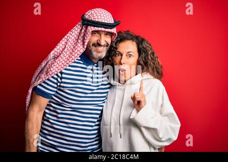 Schönes Paar im mittleren Alter mit arabischem Kopftuch und Sweatshirt, lächelnd glücklich und selbstbewusst. Mit einem Lächeln auf dem Gesicht stehend, umarmend über isoliertem Rot B. Stockfoto