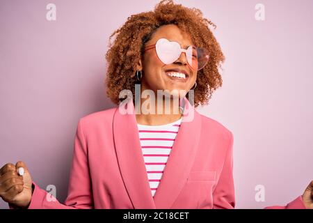 Junge afroamerikanische afro Frau mit lockigem Haar trägt lustige Herzen Sonnenbrille sehr glücklich und aufgeregt tun Gewinner Geste mit erhobenen Armen, Smilie Stockfoto