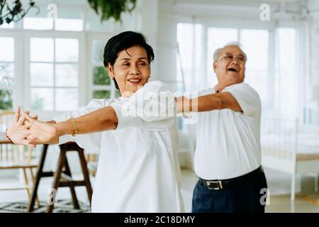 Positive ältere asiatische Mann und Frau strecken Arme und lächeln beim Tanzen zusammen in gemütlichen Raum am Wochenende Tag zu Hause Stockfoto