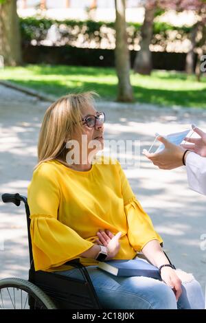 Reife Frau im Rollstuhl wird eine chirurgische Maske auf einem unfokussierten Hintergrund angeboten. Behinderung und Sicherheitskonzept. Stockfoto
