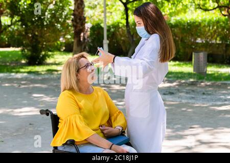 Junge weibliche Betreuerin, die eine OP-Maske auf eine lächelnde reife Frau in einem Rollstuhl auf einem unfokussierten Hintergrund setzen soll. Behinderung und Sicherheit Stockfoto