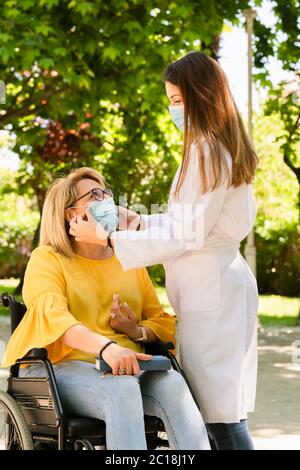 Junge weibliche Pflegekraft trägt eine chirurgische Maske Anpassung der OP-Maske einer reifen Frau in einem Rollstuhl auf einem unscharf Hintergrund. Behinderung Stockfoto