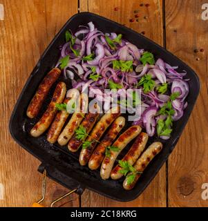 Gebratene Würstchen mit Zwiebel und Petersilie in einer Pfanne auf Holzgrund Stockfoto