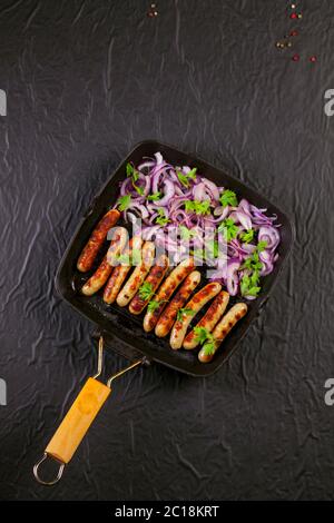 Gebratene Würstchen mit Zwiebel und Petersilie in einer Pfanne auf schwarzem Hintergrund Stockfoto