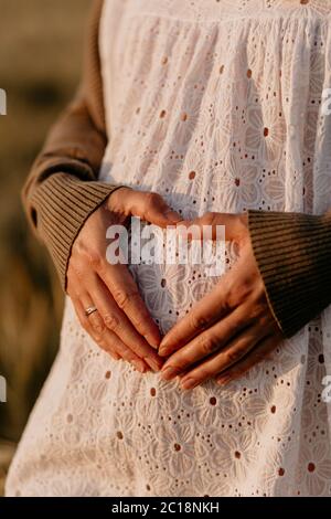 Cropped Ansicht der jungen schönen schwangeren Frau in weißem Kleid macht Herz Symbol mit Händen auf ihrem Bauch an einem sonnigen Sommertag Stockfoto